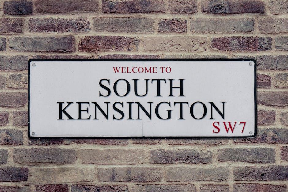 A rectangular white metal street sign mounted on a brick wall, displaying the words 'WELCOME TO SOUTH KENSINGTON' in bold black letters, with 'SW7' in smaller red font at the bottom right corner. The sign is positioned at eye level outdoors, with visible mounting screws at each corner. The brick wall behind the sign consists of reddish-brown bricks with mortar joints, providing a textured background. This image relates to house removals and relocation services available through Man and Van South Kensington, emphasizing the context of home moving or furniture transport within South Kensington's residential area.