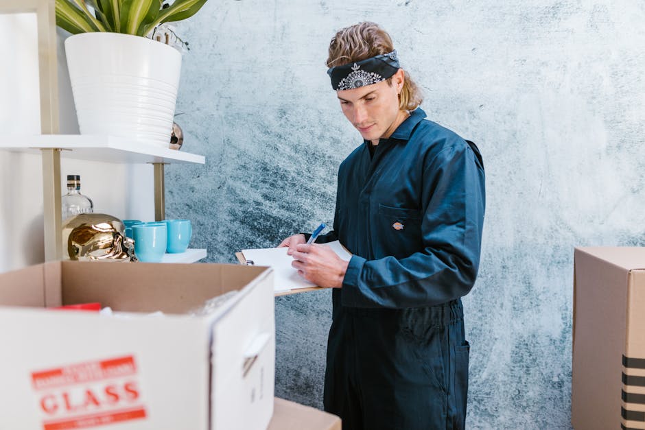 A young male mover with light brown hair, wearing a navy blue work uniform and a black bandana on his head, standing inside a room and writing on a clipboard. In front of him, there is a cardboard box filled with packing materials, including red tape and other packing supplies, on a table. Behind him, a white shelf holds a large white ceramic pot with a green plant, a transparent glass bottle, and a pair of turquoise cups. The room has a textured light grey wall with a subtle pattern and natural lighting. This scene depicts the process of home relocation, including packing and preparing household items for transport, consistent with services provided by Man and Van South Kensington during a furniture transport or moving operation.
