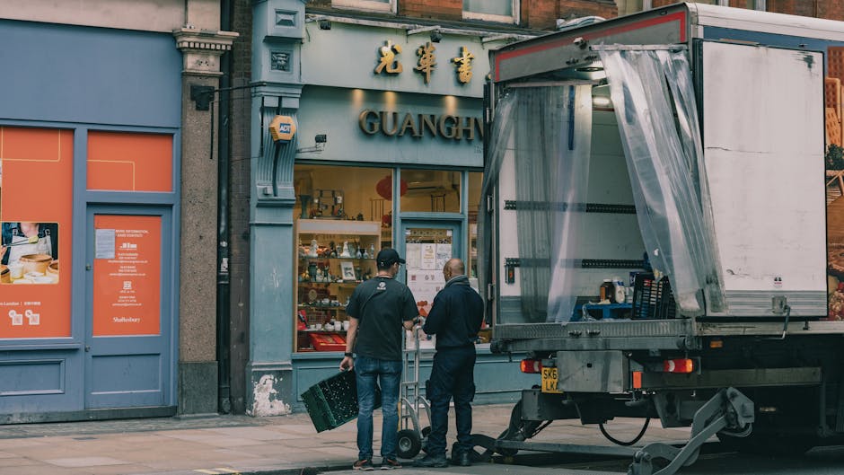 Two movers from Man and Van South Kensington are standing on the pavement in front of a storefront with Chinese characters and the word 'GUANGH' visible above the entrance, surrounded by a blue-painted facade with decorative architectural details. They are preparing to load or unload items from a large, open-sided delivery truck parked adjacent to the store. One mover is holding a black plastic crate, while the other is standing nearby, both wearing dark clothing and caps. Inside the truck's open cargo area, there are visible black and blue plastic crates and packaging materials, including bubble wrap and cardboard boxes, suggesting a home relocation or furniture transport process. The scene is set during daylight, with natural lighting illuminating the area, highlighting the urban environment typical of a South Kensington street, supporting a professional removals service focused on packing and moving tasks.