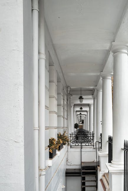 A long, narrow exterior corridor of a residential building featuring white-painted walls, columns, and decorative brackets. Potted plants with flowers are placed along the left side, and black metal outdoor lanterns hang at intervals from the ceiling. A black metal railing borders a staircase that leads down to a lower level. In the foreground, a man in casual clothing is partially visible, holding or securing a large cardboard box wrapped in plastic with protective moving blankets. The scene appears to capture a home relocation or furniture transport process taking place on a bright day, with natural light illuminating the area and suggesting an active loading or unloading activity typical of end-of-tenancy removals, consistent with services offered by Man and Van South Kensington.