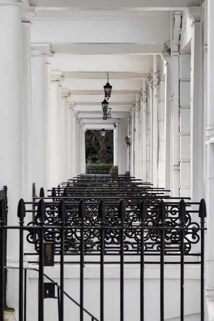 A long, white, covered porch of a residential building featuring evenly spaced columns supporting the roof, with black wrought iron decorative railings at the front of the porch. The porch ceiling has hanging lantern-style light fixtures, and the floor appears to be made of concrete or stone slabs. A large tree is visible in the background beyond the porch, with some greenery and bushes. This setting suggests an exterior view of a house in South Kensington, possibly part of a property prepared for a home relocation or removal process, with the front entrance and decorative elements clearly visible, aligned with house removal services provided by Man and Van South Kensington.