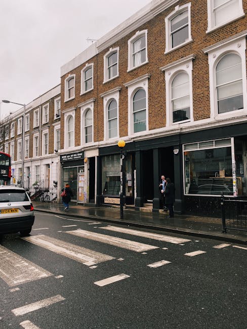 A street scene outside a three-storey brick building on Earls Court Road in South Kensington, with large windows and a black storefront. The pavement is wet, indicating recent rain, and there are pedestrians, some wearing coats and holding umbrellas, walking along the sidewalk. A white car is parked alongside the curb, and a red double-decker bus is visible further down the street. In front of the building, a man in dark clothing stands next to a black van, part of a home relocation or furniture transport process facilitated by Man and Van South Kensington. Nearby, a person is carrying a cardboard box, surrounded by packing materials such as bubble wrap and boxes, suggesting ongoing packing and moving activities. The scene captures the typical urban environment associated with house removals in South Kensington, with the loading area busy with loading or unloading furniture, boxes, and packing supplies related to a recent or upcoming move, aligned with professional removals services.