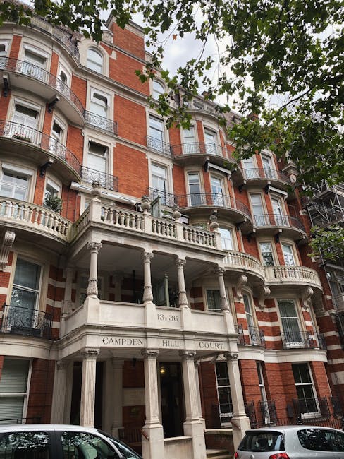 The image shows the façade of a multi-storey red brick residential building with ornate white stone columns and balustrades at the entrance, identified as Campden Hill Court. Large windows with black metal railings are visible on several floors, and a canopy extends over the main doorway. The building is surrounded by leafy green trees, some branches hanging in the upper part of the image. In the foreground, parked cars are visible along the pavement. The scene takes place during daylight with natural light illuminating the structure. The photograph captures the exterior details of the building, which is relevant to house removals and furniture transport services, as provided by Man and Van South Kensington, highlighting the type of property involved in home relocation or moving logistics in South Kensington.