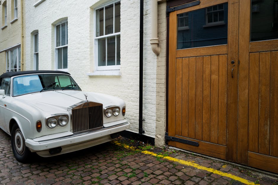 A white vintage car with a black soft top is parked on a cobblestone driveway next to a white brick building with two large sash windows. The car's front grille and headlights are clearly visible, and a hood ornament is mounted on the front. Behind the car, there is a wooden sliding gate, slightly open, leading to an interior space. The scene is lit with natural daylight, and the driveway shows signs of weathering, with moss and dirt along the edges. Slightly visible near the building's base are yellow painted lines indicating parking boundaries. The setting reflects a typical London residential area suitable for house removals, and Man and Van South Kensington regularly handles furniture transport and packing during home relocation projects, often working in tight urban environments like this one.