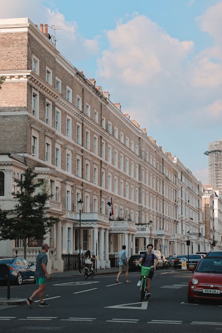 Image of a row of elegant, multi-storey Victorian terraced houses in South Kensington, London, with detailed brickwork, sash windows, and decorative white window frames. The street scene features a clear blue sky with some clouds, and parked cars are visible along the pavement. Pedestrians, including a man on a bicycle and others walking, are present on the asphalt road. The image captures the environment typical of a residential area suitable for house removals and furniture transport, with the street ready for the loading process. Man and Van South Kensington operates in this area, providing professional removals services involving packing, loading, and transporting household furniture and belongings, often requiring parking permits and adherence to council rules for parking and access.