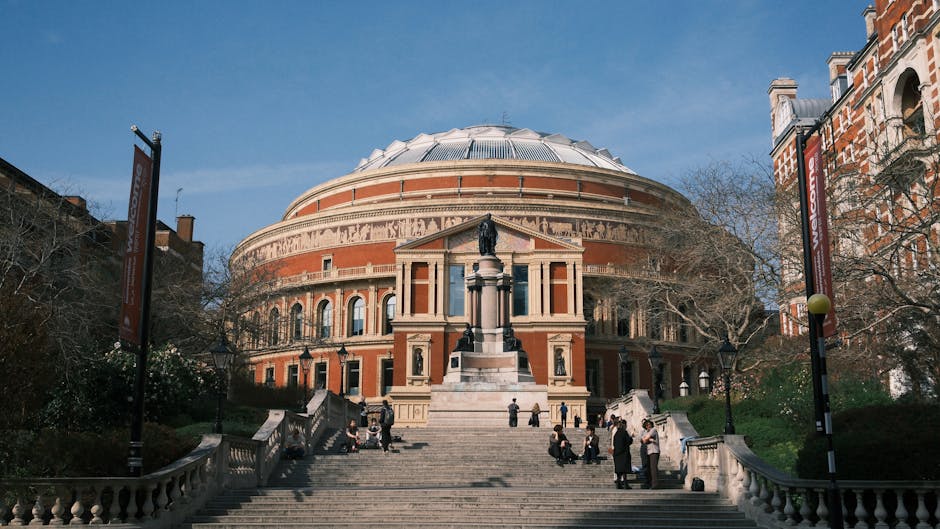 Exterior view of the Royal Albert Hall in South Kensington, showcasing its iconic round, domed roof and red-brick facade with decorative arches and columns. The building is accessed by a broad stone staircase leading up to the entrance, where a statue of a historical figure stands on a tall pedestal near the top of the stairs. Surrounding the staircase are metal railings, some people are seated on the steps or standing engaged in conversation. To the left and right, there are trees with bare branches and lampposts lining the pathway, indicating a clear, sunny day. The scene captures a moment of activity during a home relocation or furniture transport operation, with a van parked nearby, possibly involved in packing and moving processes related to a house move or event, with Man and Van South Kensington providing a professional service for the area.