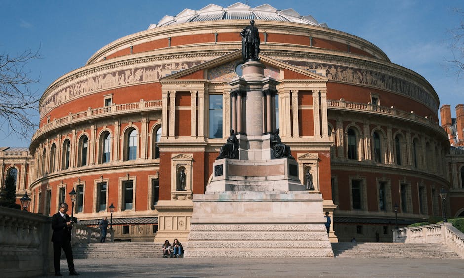 Exterior view of the Royal Albert Hall in South Kensington, showcasing its iconic round, domed roof and red-brick facade with decorative arches and columns. The building is accessed by a broad stone staircase leading up to the entrance, where a statue of a historical figure stands on a tall pedestal near the top of the stairs. Surrounding the staircase are metal railings, some people are seated on the steps or standing engaged in conversation. To the left and right, there are trees with bare branches and lampposts lining the pathway, indicating a clear, sunny day. The scene captures a moment of activity during a home relocation or furniture transport operation, with a van parked nearby, possibly involved in packing and moving processes related to a house move or event, with Man and Van South Kensington providing a professional service for the area.