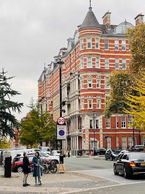 Photograph of a busy urban street scene in South Kensington, showing a large, historic, red-brick building with white stone detailing and multiple bay windows. The building features a rounded turret w