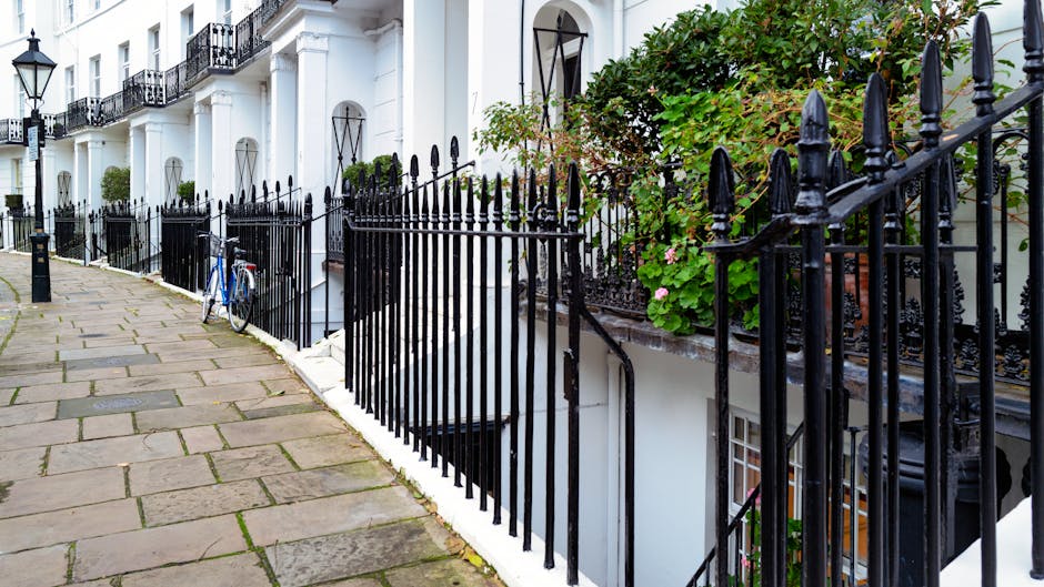 A narrow London street with a paved sidewalk composed of large rectangular stone slabs, running alongside row houses with white facades and black wrought iron railings. A blue bicycle is parked against the railing near a basement entrance, which is partly visible beneath a staircase leading down from a residential building. The stairs and entrance are enclosed by the black metal fencing that extends along the street. Some green foliage and flowering plants grow above the railing, adding a touch of greenery to the scene. The lighting appears natural, indicating daytime, with moderate shadows cast by the railings and plants. The image captures a typical urban setting suited for house removals and furniture transport services, emphasizing the importance of careful logistics when moving items through such confined outdoor environments as part of a home relocation in South Kensington, as highlighted on the service page for Gloucester Road flats.