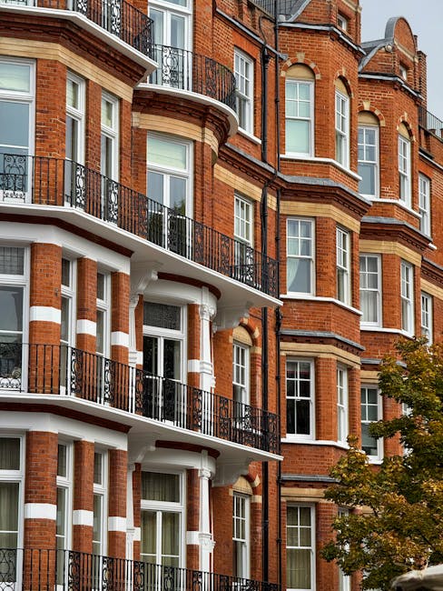 A multi-storey red brick residential building with white decorative accents and black wrought iron balconies is visible in the image. Several large windows on each floor allow natural light into the interior apartments. In the foreground, a tree with green leaves partially obscures the lower part of the building. The building appears to be well-maintained, with clean brickwork and secure balcony railings. This scene is characteristic of typical London residential architecture found in South Kensington, supporting the context of house removals and furniture transport services provided by Man and Van South Kensington during home relocation or packing and moving processes. The natural daylight highlights the building's architectural details, emphasizing its height and urban setting, suitable for the visual description of a property involved in a professional removals service for Gloucester Road flats.